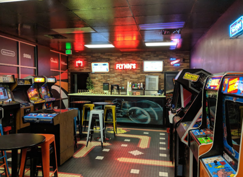 A retro arcade with colorful neon lights, featuring classic arcade machines and a bar area in the background.