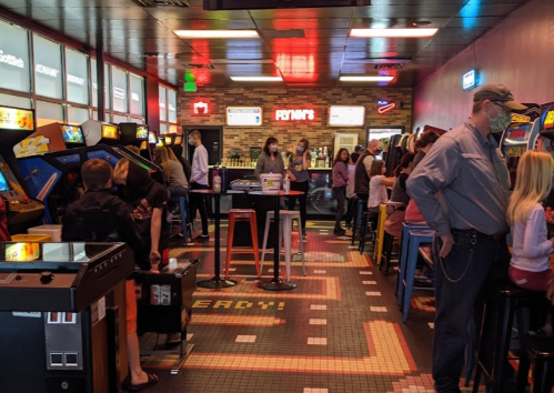 A lively arcade filled with people playing games, colorful lights, and retro machines lining the walls.
