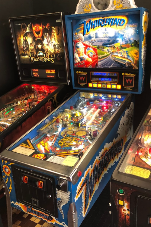 A colorful pinball machine titled "Whirlwind" with vibrant artwork and flashing lights, surrounded by other machines.