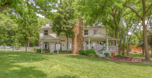 A charming two-story house surrounded by lush greenery and trees, featuring a stone chimney and a welcoming porch.