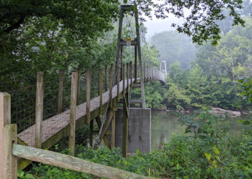 A wooden suspension bridge spans a river, surrounded by lush greenery and misty trees.