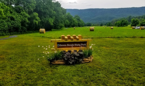 Sign for Seven Bends State Park in a grassy field with hay bales and mountains in the background.