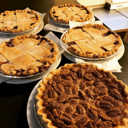 A display of five freshly baked pies, including fruit-filled and pecan varieties, on white plates.
