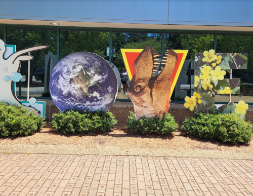 Colorful letters spelling "LOVE" with Earth, a dinosaur, and flowers, set against a glass building and greenery.