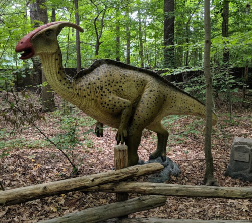 A life-sized dinosaur statue in a wooded area, surrounded by trees and fallen leaves, with a wooden fence nearby.