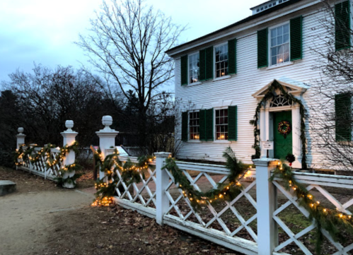 A white house with green shutters, decorated with garlands and lights for the holidays, surrounded by bare trees.