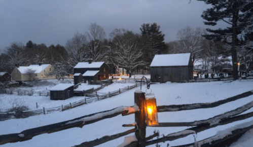 A snowy landscape with rustic buildings and a glowing lantern, surrounded by trees under a cloudy sky.