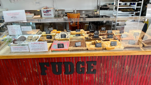 A display case filled with various types of fudge, labeled with names, in a cozy shop setting.