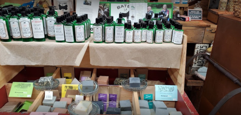 A display of green bottles of goat lotion and various colorful soap bars in a rustic shop setting.