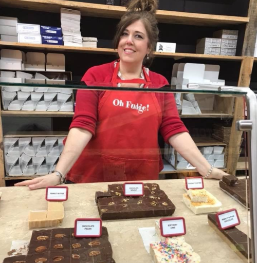 A smiling woman in a red apron stands behind a display of various fudge flavors in a candy shop.