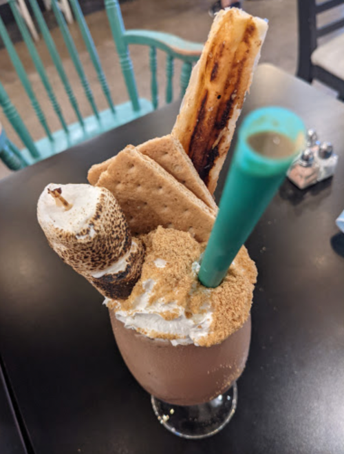 A decadent dessert drink topped with whipped cream, a toasted marshmallow, graham crackers, and a chocolate stick.