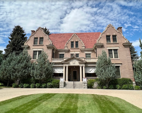 A large, historic mansion with a red roof, surrounded by green trees and a well-manicured lawn under a cloudy sky.