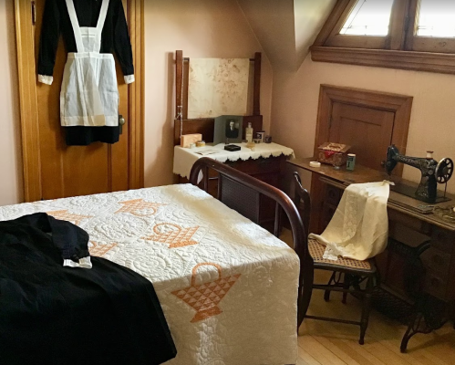 A vintage bedroom with a quilted bed, sewing machine, and a dress hanging on the wall. Warm, nostalgic atmosphere.