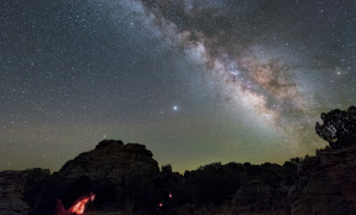 A stunning night sky filled with stars and the Milky Way, framed by rocky formations and a hint of glowing light below.