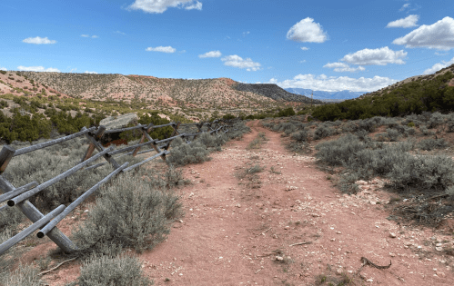 A dirt path winds through a desert landscape with shrubs and a wooden fence under a blue sky with clouds.