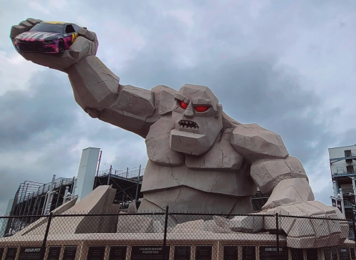 A giant stone monster with red eyes holds a race car in its hand, set against a cloudy sky.