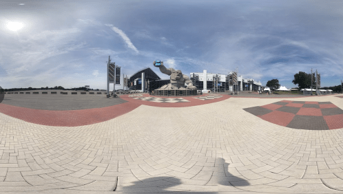 A panoramic view of a plaza featuring a large sculpture, checkered patterns, and a modern building under a blue sky.