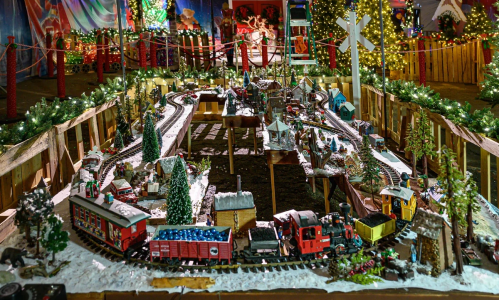 A festive model train set with miniature buildings, trees, and decorations, surrounded by holiday lights.