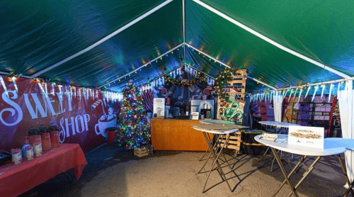 A festive tent decorated with lights, a Christmas tree, and tables for a sweet shop event.