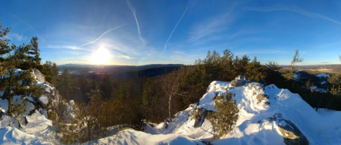 A panoramic view of a snowy landscape with trees and mountains under a bright blue sky and setting sun.