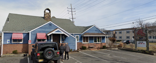 A blue and brick building with a stone chimney, featuring a sign and parked cars outside on a clear day.