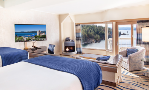 Cozy hotel room with two beds, a TV, and a scenic view of a lake and forest through large windows.