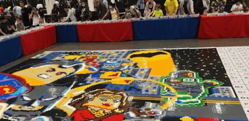 A large mosaic made of colorful blocks depicts superheroes, with people assembling it in a crowded venue.