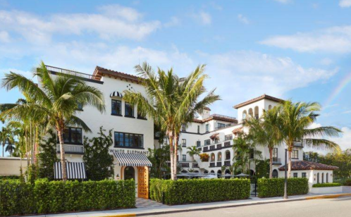 A stylish hotel with white walls, palm trees, and a blue sky, featuring balconies and lush greenery in the foreground.