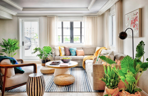Cozy living room with a beige sofa, colorful cushions, plants, a round coffee table, and large windows letting in natural light.