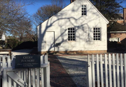 A historic white building with a sign reading "Geddy Foundry" in a snowy landscape, surrounded by a white picket fence.