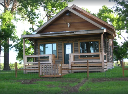 A cozy wooden cabin with a porch, surrounded by green trees and a fenced yard.