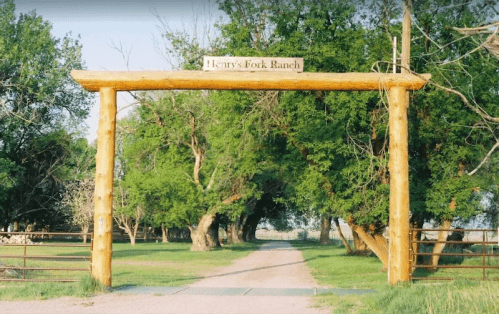 Wooden entrance gate to Henry's Fork Ranch, flanked by green trees and a gravel driveway leading into the ranch.