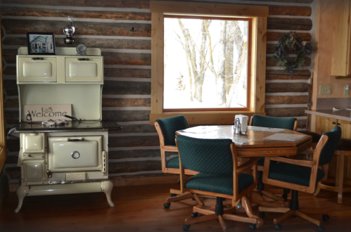 A cozy log cabin interior featuring a vintage stove and a wooden dining table with green chairs by a large window.