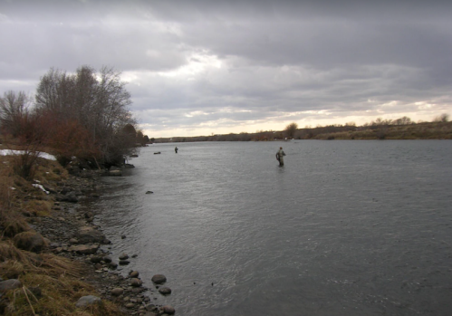 Two anglers wade in a river under a cloudy sky, surrounded by trees and rocky banks.