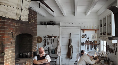 A historic workshop with two men working, surrounded by tools and a brick fireplace, showcasing a rustic interior.