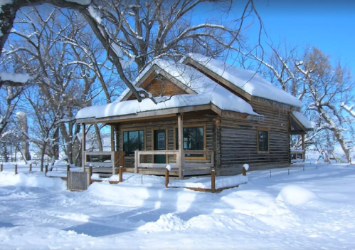 A cozy wooden cabin covered in snow, surrounded by trees under a clear blue sky.