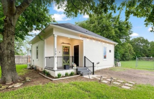 A charming white house with a front porch, surrounded by greenery and a gravel pathway.