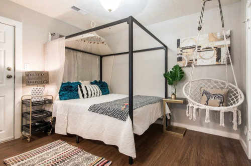 Cozy bedroom with a four-poster bed, hanging chair, and decorative plants, featuring warm wood flooring and soft textiles.