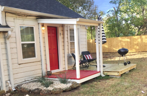 A cozy house entrance with a red door, porch, and grill, surrounded by greenery and a wooden fence.