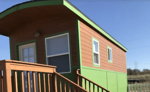 A small, colorful house with orange and green siding, featuring a wooden deck and multiple windows.