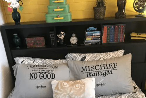 Cozy bedroom with Harry Potter-themed pillows, books, and decorative items on a black shelf against a yellow wall.