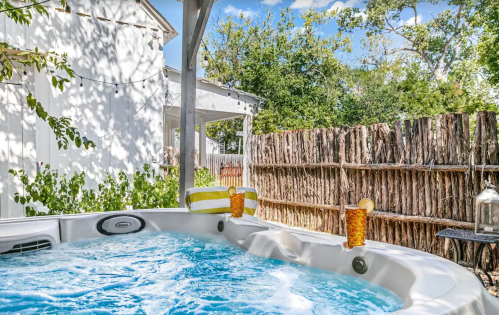 A hot tub with bubbling water, surrounded by greenery and a rustic fence, with two drinks on the edge.