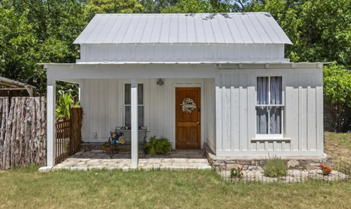 A charming white cottage with a metal roof, front porch, and garden, surrounded by greenery.