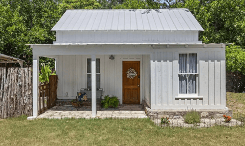 A charming white cottage with a metal roof, front porch, and garden, surrounded by greenery.
