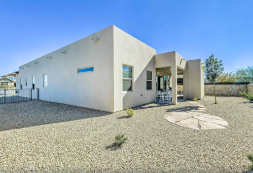 A modern, minimalist house with a gravel yard and patio area, surrounded by low landscaping under a clear blue sky.