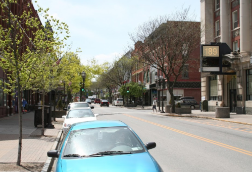 A quiet street lined with trees and parked cars, featuring buildings and a sign displaying "189."