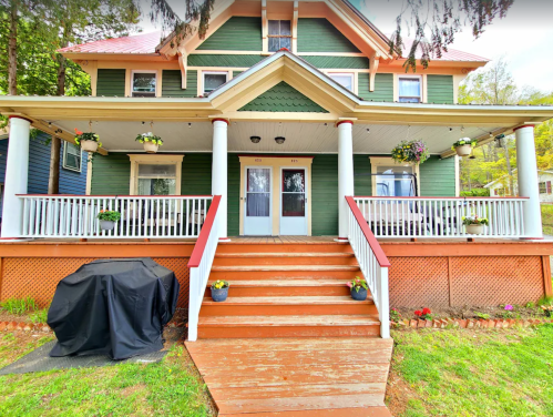 A green two-story house with a large porch, white railings, and flower pots, featuring steps leading to double doors.