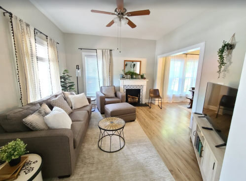 Cozy living room with a gray sofa, armchair, coffee table, and a fireplace, featuring light curtains and wooden floors.