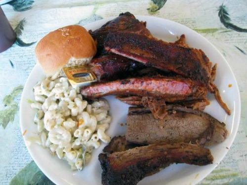 A plate of barbecue ribs, brisket, macaroni salad, and a bun with butter on a patterned tablecloth.