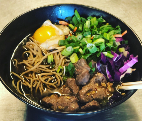 A bowl of ramen with beef, a poached egg, green onions, and purple cabbage on top of soba noodles in broth.